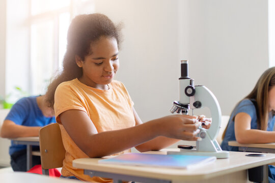 Black schoolgirl adjusting microscope during science lesson in bright classroom, preparing for an experiment while diverse classmates work at desks in the background