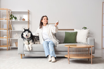 Pet ownership and home comfort, woman relaxing on sofa with husky dog using air conditioner remote control in modern living room interior © Pixel-Shot