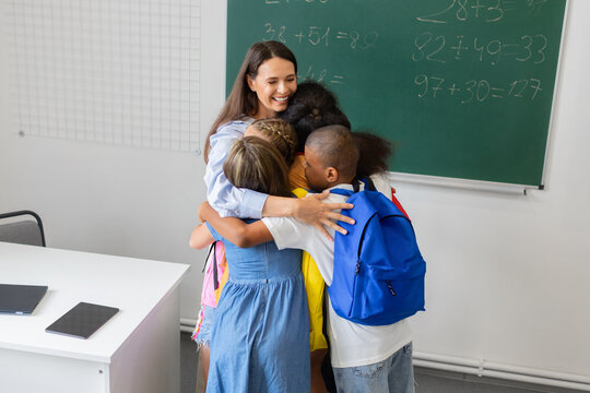 Smiling female teacher sharing warm group hug with diverse schoolchildren wearing backpacks in a classroom, showing trust, support, inclusion and positive relationships