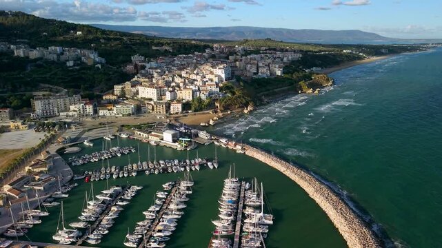 Rodi Garganico Puglia Italy 4k drone view of marina yachts at sunrise. Backward pullback flyaway of semi circular breakwater and whitewashed coastal town b-roll scenic establishing video footage