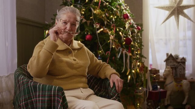 Portrait of senior Caucasian woman putting on eyeglasses and smiling while sitting near Christmas tree. Perfect for holiday marketing, senior care ads, and festive family greeting promotions