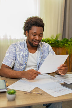 African American young man on home table sorts documents into piles and finds needed page with satisfaction. Black guy on home table checks papers and nods after finding important document for work.