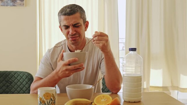 Adult man with funny skeptic and confused facial expression holding spoon and tasting weird breakfast cereal while sitting at the kitchen dining table