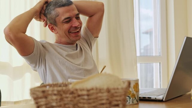 Satisfied happy male freelancer leaning back with hands behind head celebrating finished remote project on laptop sitting at the kitchen table in home office