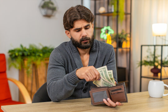Indian man at home opens wallet sees small dollar cash frowns worries about bills debt low salary plans budget. Arabian guy at desk counts coins checks costs plans reduce spend records poverty risk.