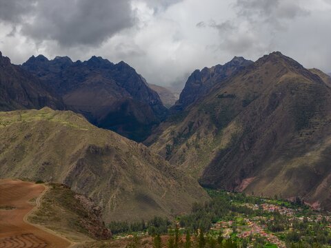Aerial view of the Andes mountains and the town of Ollantaytambo nestled in the Sacred Valley with rugged slopes, green vegetation, and cloudy skies in Ollantaytambo, Cusco, Peru.