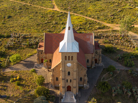 Aerial view of a historic stone church with a tall steeple surrounded by arid landscape, dirt paths, and native quiver trees in Oudtshoorn, Western Cape, South Africa.