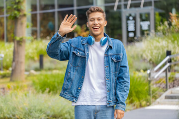 Young man waving hello with grin while walking in city street and greeting camera warmly. Adult guy tourist giving welcome gesture outdoors with relaxed mood and cheerful face during calm urban walk. © Andrii Iemelianenko