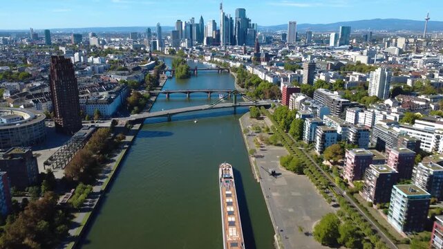 Aerial flight along the Main River toward Frankfurt skyline with a river cruise ship and multiple city bridges. Filmed September 25, 2026.