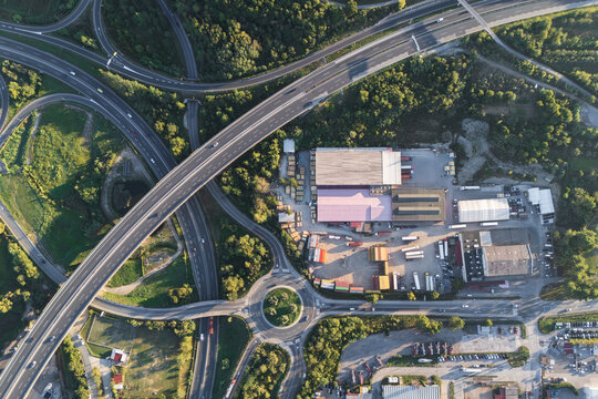 Aerial view of the Sermin industrial area featuring large warehouses, logistics facilities with parked trucks, and a complex highway interchange with a roundabout in Koper, Koper, Slovenia.