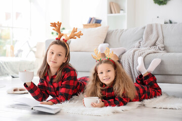 Obraz premium Cute sisters in reindeer horns with cocoa reading story at home on winter day