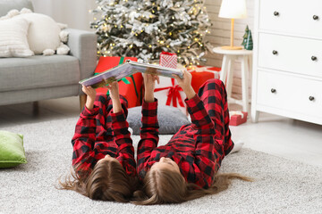 Obraz premium Cute little girls in checkered pajamas with book reading Christmas story at home
