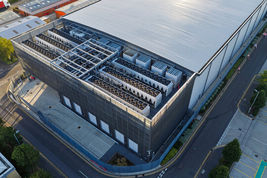 Aerial view of a modern data center facility featuring industrial cooling units on the roof, surrounded by asphalt parking areas and roads in the United Kingdom.