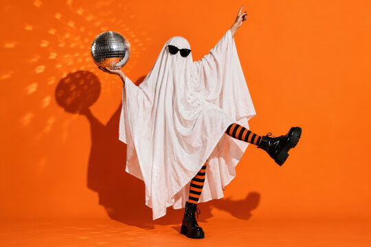 Sheet ghost in sunglasses holding up a mirror disco ball mid kick, striped tights and platform boots. Funny Halloween disco party in a studio shot
