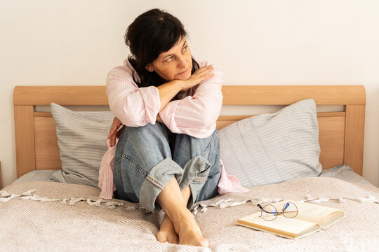 Mature woman in jeans and pink shirt sits on bed, arms wrapped around her knees, looking pensive and gazing off to side, book and glasses lie nearby. Measured pace of life, mindful leisure
