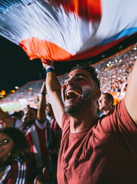 Paraguay football fan with a painted face cheering and holding a flag overhead in a crowded stadium for sports and national pride