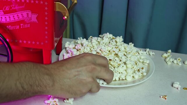 A close-up shows a hand grabbing fresh popcorn from a plate beside a red retro popcorn machine. The indoor movie night setting features spilled kernels and moody blue lighting.