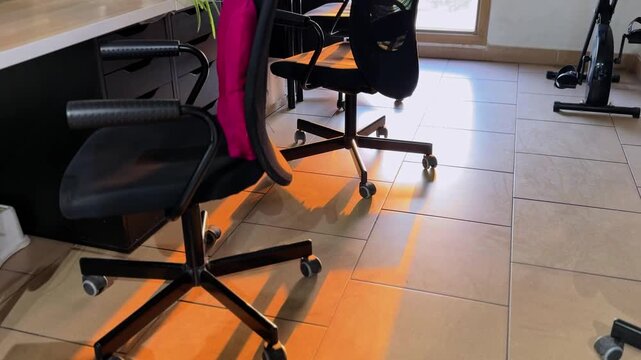 Golden sunset light streams across a tiled floor, highlighting empty black swivel chairs in a modern home office. An exercise bike rests in the quiet background.