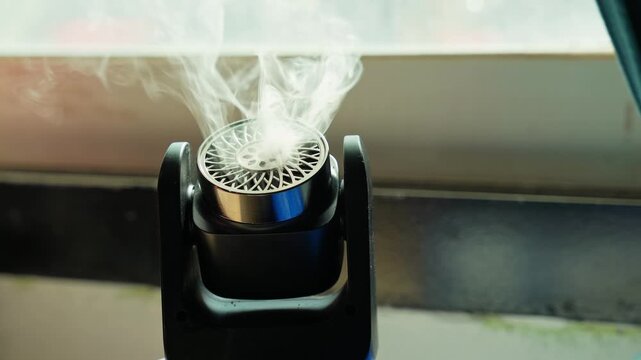 A close-up view of a small black electronic diffuser emitting white smoke indoors. The portable device rests near a bright window with natural daylight illuminating the rising vapor.