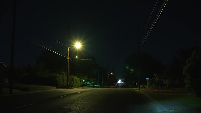 Empty dark street at night illuminated by sparse streetlights among houses, trees and power lines, creating a moody and tense atmosphere
