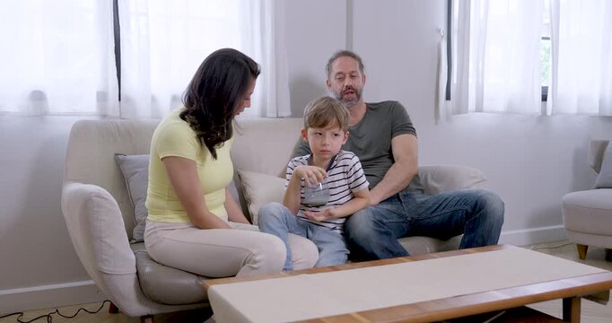 Young son drinks milk between Asian mother and Caucasian father, then rises from sofa as father supports movement, combining family conversation, attentive childcare, and everyday bonding at home.
