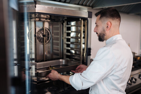 Chef works in a busy kitchen preparing food using an oven and taking out a tray with freshly cooked items