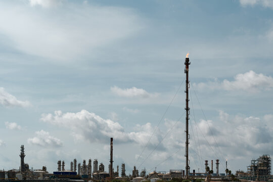Tall industrial gas flare stack with guy wires burning off excess hydrocarbons at large crude oil refinery and petrochemical processing plant with fractionating columns under cloudy sky.