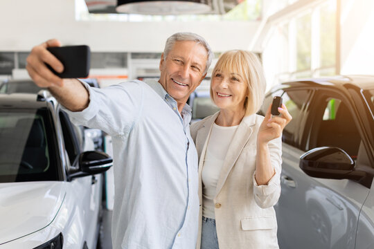 Cheerful senior Caucasian couple taking selfie inside car dealership showroom while the woman displays vehicle key, celebrating their successful new automobile purchase together