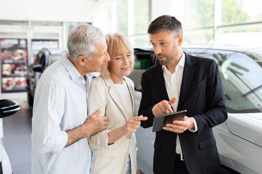 Professional car salesperson using tablet to explain vehicle details to senior Caucasian couple in a dealership showroom as they review options before buying