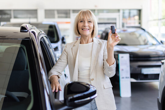 Smiling senior Caucasian woman standing beside vehicle in dealership showroom and holds up the car key, representing successful purchase, delivery, and ownership