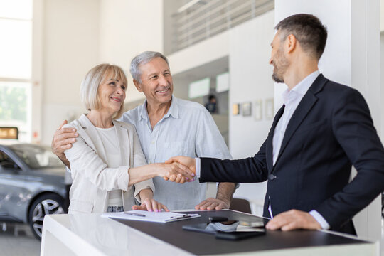 Senior Caucasian woman shaking hands with car salesperson at dealership desk while her partner stands beside her, celebrating a successful vehicle purchase agreement