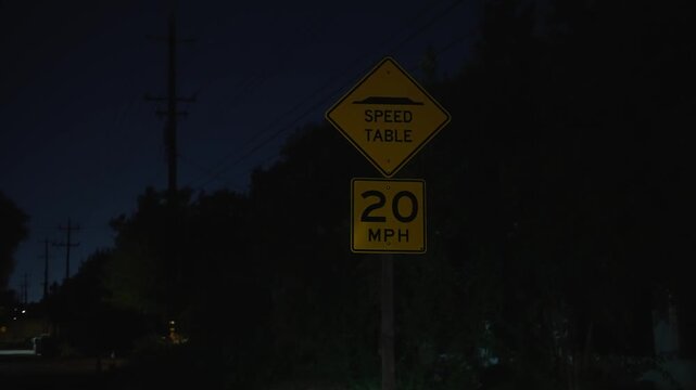 Speed warning sign stands beside a dark road at night, indicating a speed table and recommended speed of 20 miles per hour