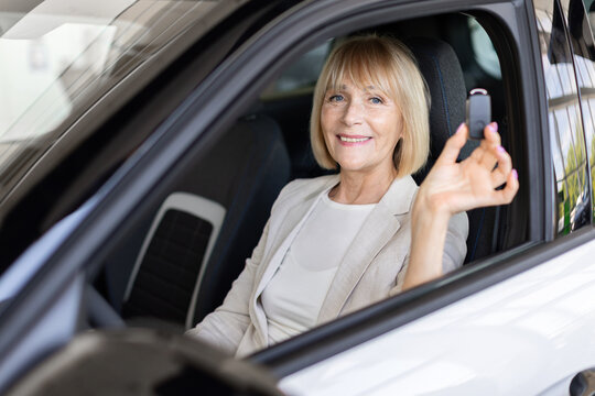 Smiling senior Caucasian woman sitting inside new vehicle at dealership showroom and displays the car key, celebrating successful purchase and automobile ownership