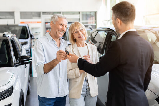 Happy senior Caucasian couple receiving car key from salesperson in a bright dealership showroom, marking the successful handover and completion of their vehicle purchase