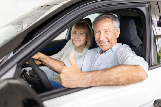 Smiling senior caucasian couple sitting inside new car at dealership showroom as the man gives thumbs up, expressing approval and satisfaction with the vehicle