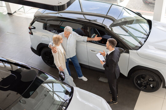 Senior Caucasian couple discusses a vehicle with professional salesperson beside new SUV in dealership showroom, receiving advice while comparing options before purchase