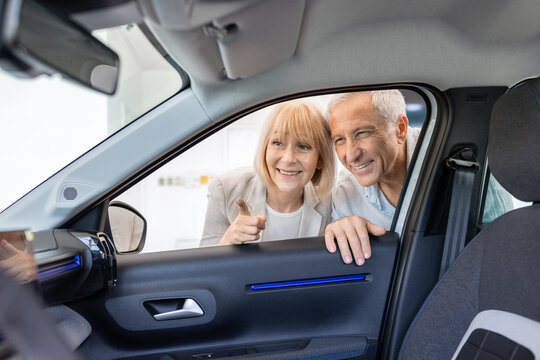 Senior Caucasian couple looking through an open car door at the vehicle interior in a dealership showroom, pointing out cabin details while evaluating features before purchase