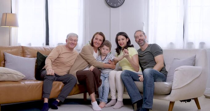 Young son sits between Asian mother and Caucasian father as senior grandparents lean close, smile, and wave toward camera, creating cheerful greeting and warm multigenerational family bonding at home.
