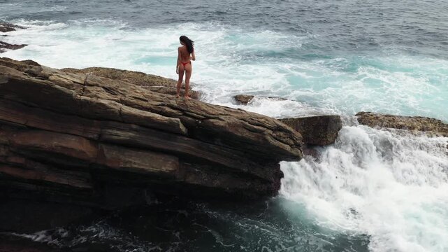 Woman in Red Bikini Standing on Coastal Rocks by Crashing Ocean Waves