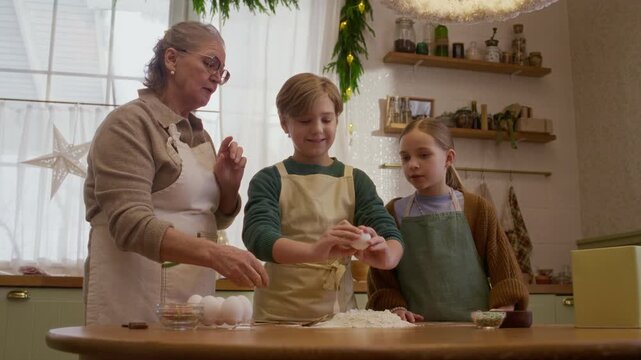 Caucasian senior woman teaching grandson cracking egg into flour beside granddaughter in kitchen. Footage suited for family baking campaigns, holiday cooking commercials, culinary education