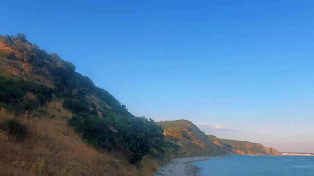 Late afternoon sun illuminates dry grassy hills sloping steeply toward a calm sea shoreline along Saros Bay, Edirne, Turkey.