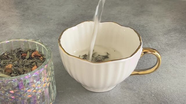 Hot water pouring into white elegant cup with gold rim filled with loose leaf herbal tea leaves on grey concrete background. Tea leaves swirling and blooming in slow motion, cozy aesthetic.
