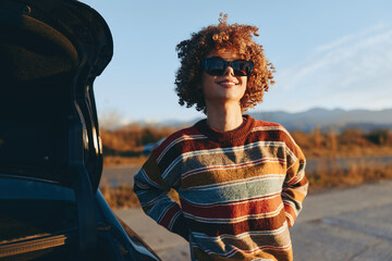 Obraz premium Woman with curly hair smiling outdoors, wearing striped sweater, standing near car on road, sunlight golden hour, relaxed lifestyle mood, soft breeze, warm natural colors, distant hills
