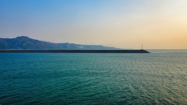 Scenic long stone breakwater jetty stretching into the blue sea under a golden sunset sky with mountain range in the distance, tranquil coastal landscape for travel and nature concepts.
