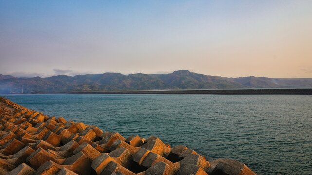 Rows of concrete breakwater blocks or tetrapods protecting the coastline with a scenic view of the calm blue sea and majestic mountain range under a soft sunset sky at a harbor or coastal bay.