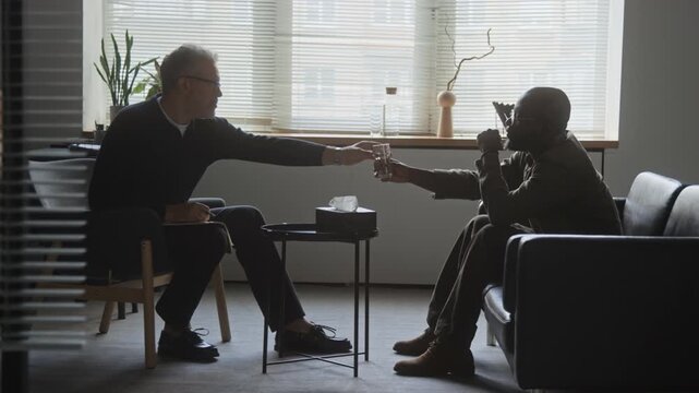 Middle aged Caucasian therapist giving glass of water to African American male client during psychotherapy session. Suitable for mental health campaigns, psychological counseling, therapy adverts