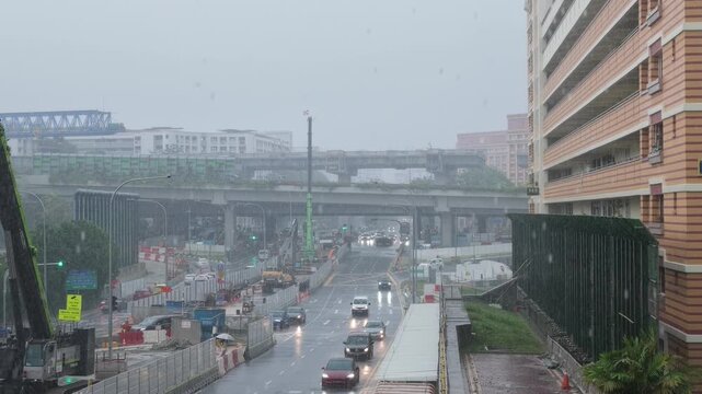 Real-time footage of a rainy day in the city, capturing active vehicle traffic on wet streets, residential houses, and an ongoing bridge construction site under a gray sky.