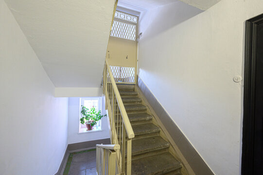 Narrow residential staircase with concrete steps, pale yellow railing, white walls, window with potted plant, upper landing with metal-grid window