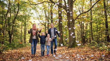 Fototapeta premium Happy Family Walking in Autumn Forest with Paper Cranes and Leaves Flying. Concept of origami birds, falling leaves, kids, parents, outdoor activity.