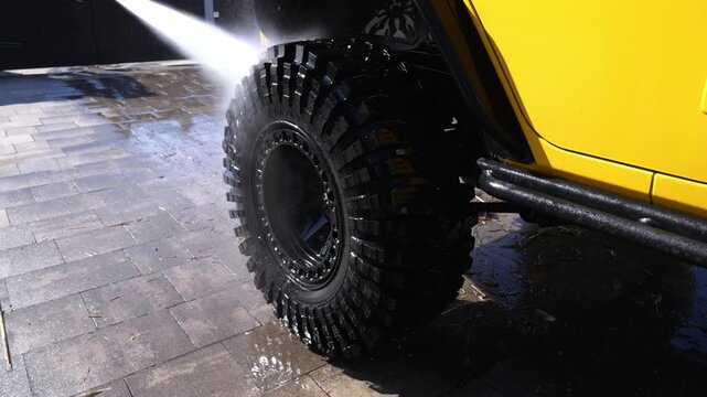 High-pressure water jets wash off dirt from a rugged off-road tire. Bright yellow vehicle stands ready for cleaning. Water sprays forcefully, clearing mud and grime